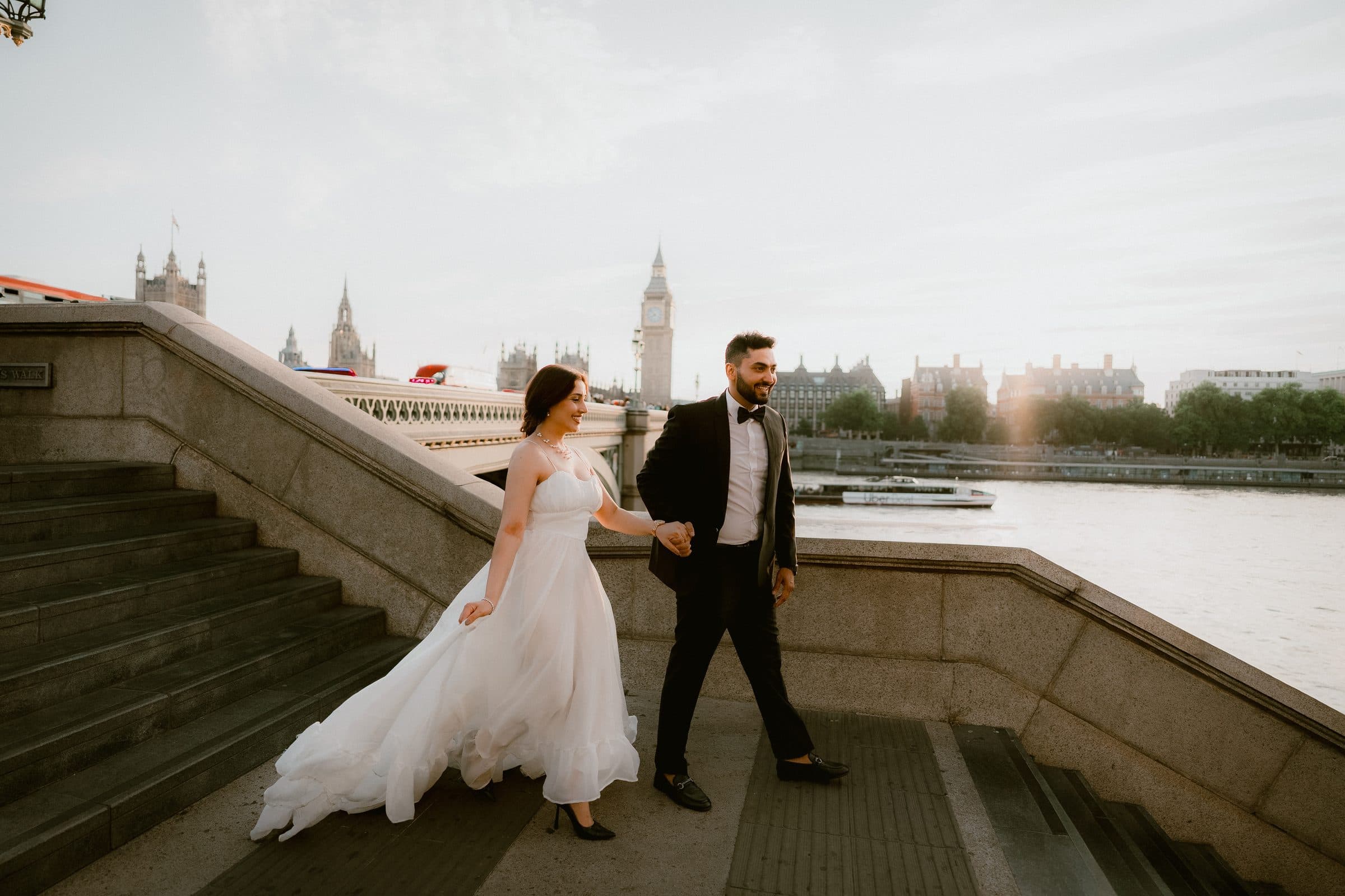 A couple walking down stone steps beside the Thames with Big Ben across the water during a London engagement photoshoot