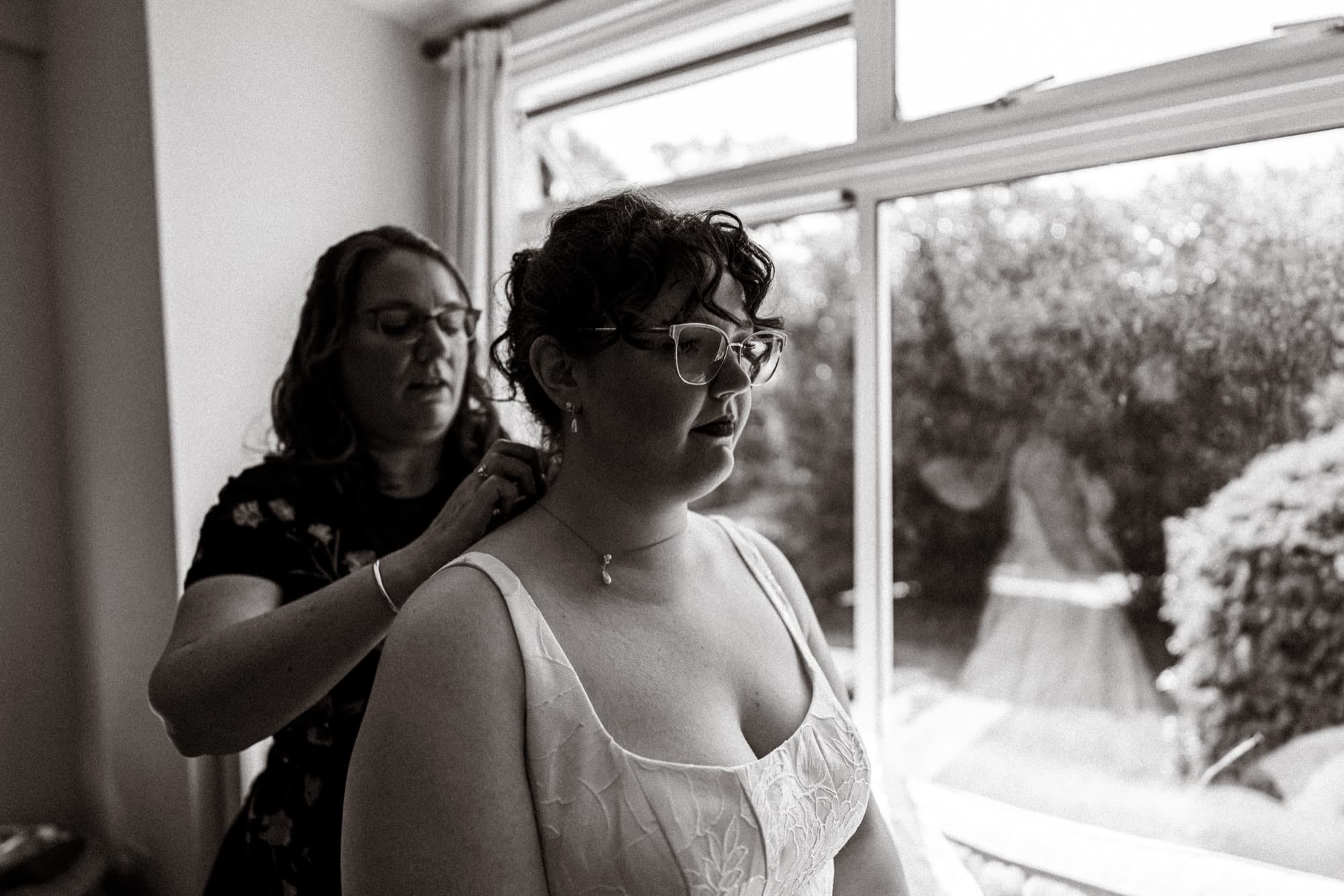 Black and white photograph of the bride having her dress fastened by the window, her gown reflected in the glass, before the All Souls, Langham Place wedding