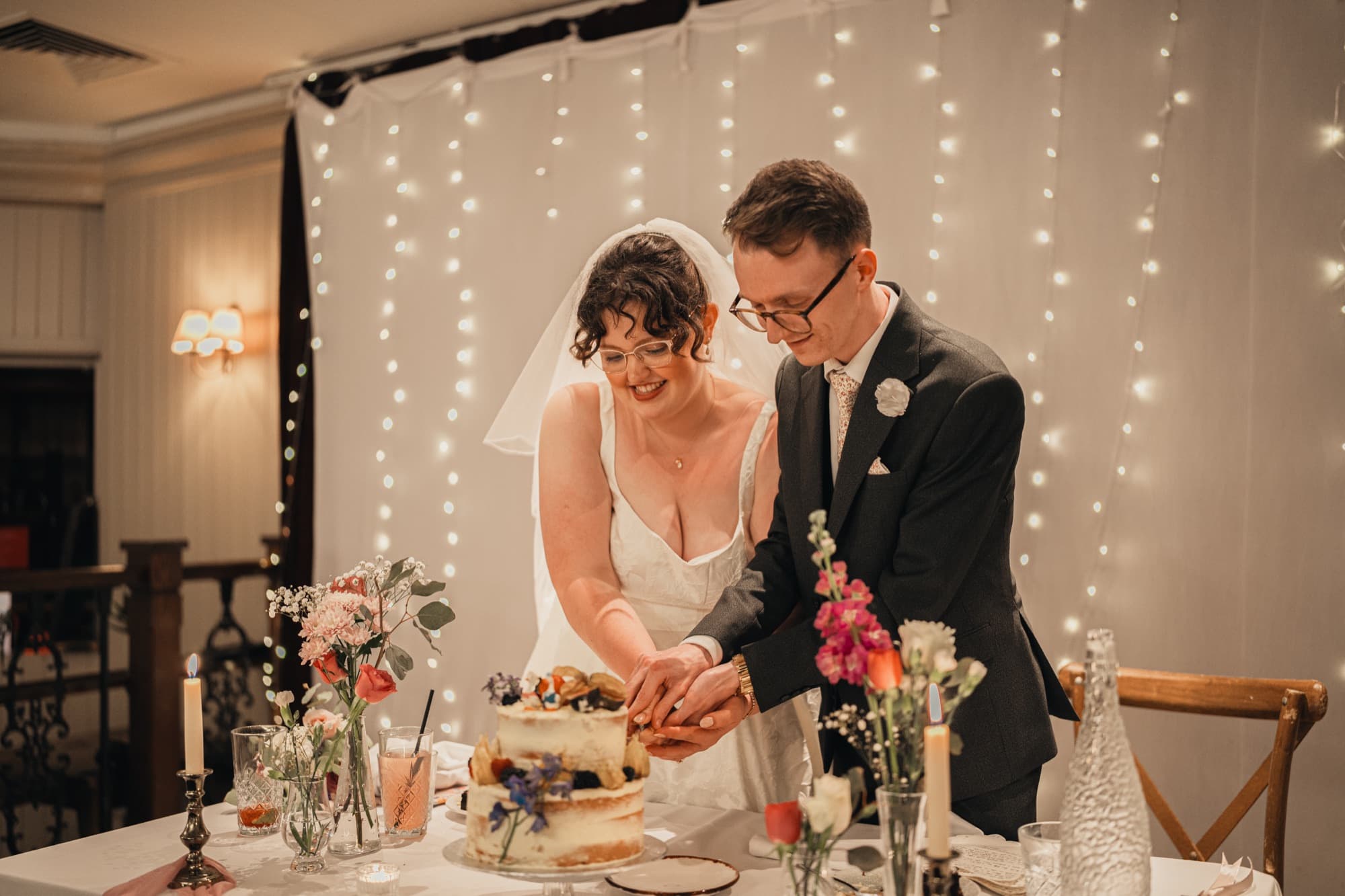 Dylan and Arianna cutting their wedding cake beneath a backdrop of fairy lights at their reception