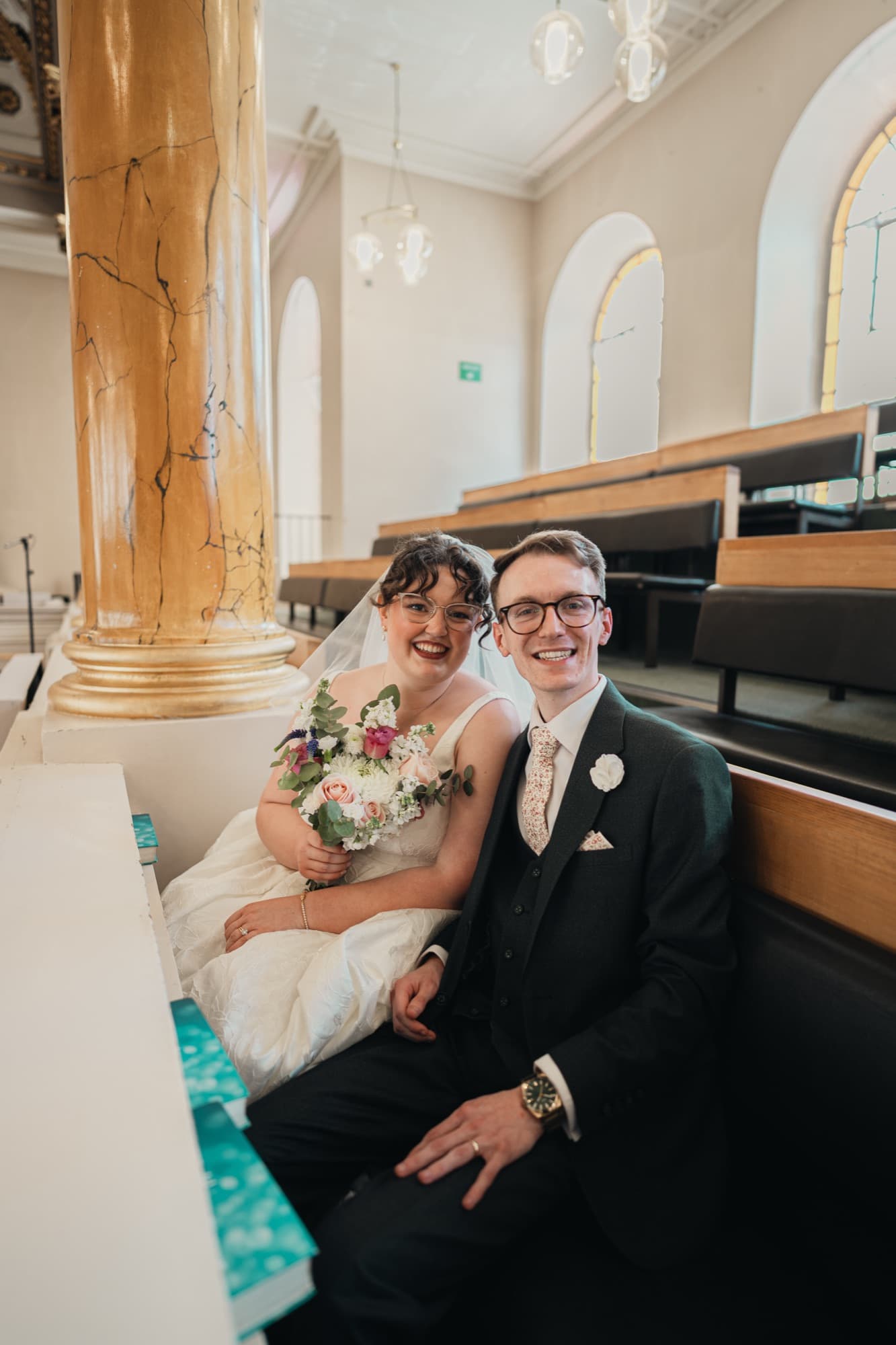 Relaxed portrait of Dylan and Arianna seated in a pew beneath a gilded column at All Souls, Langham Place