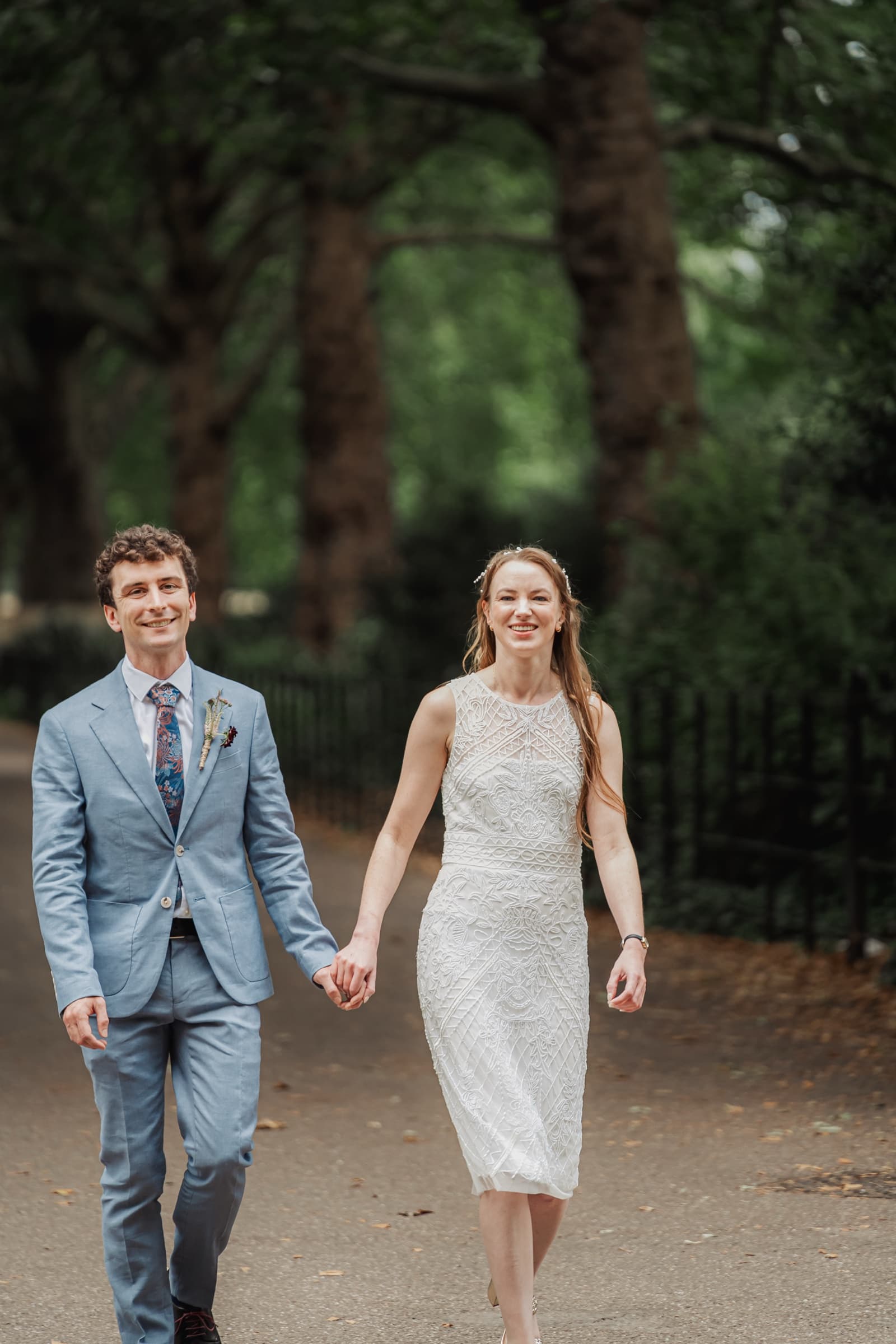 Bride and groom walk hand in hand along a tree-lined avenue in Battersea Park