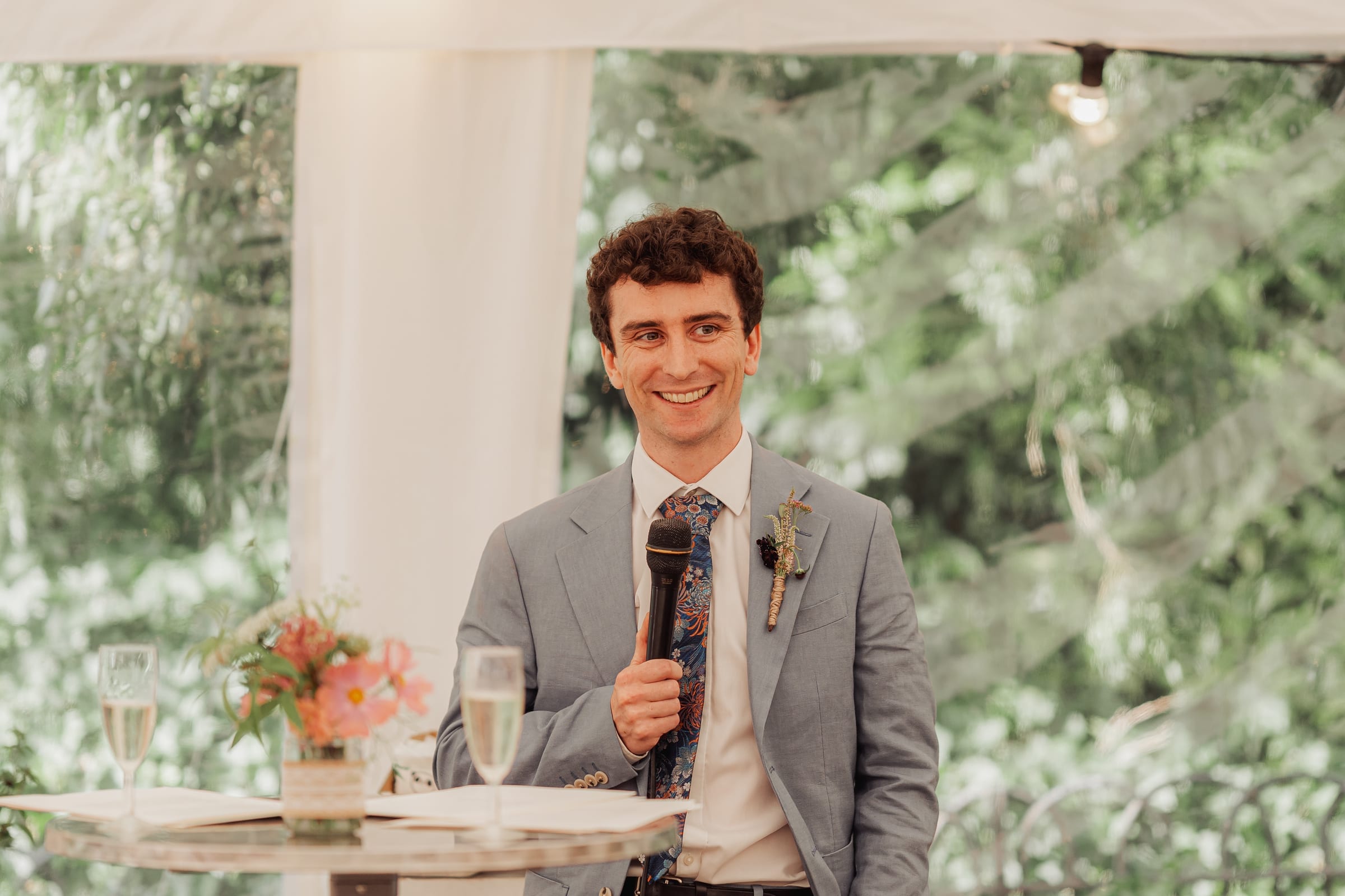 A speaker smiling mid-speech with a microphone at a garden celebration
