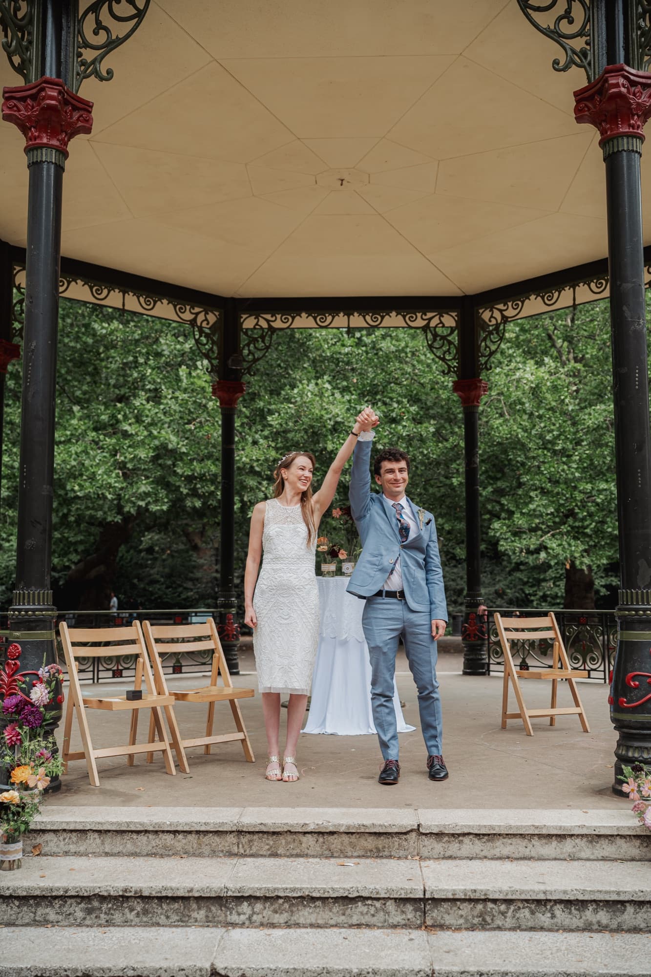 Newly married couple raise their clasped hands in triumph on the bandstand steps at Battersea Park, both beaming